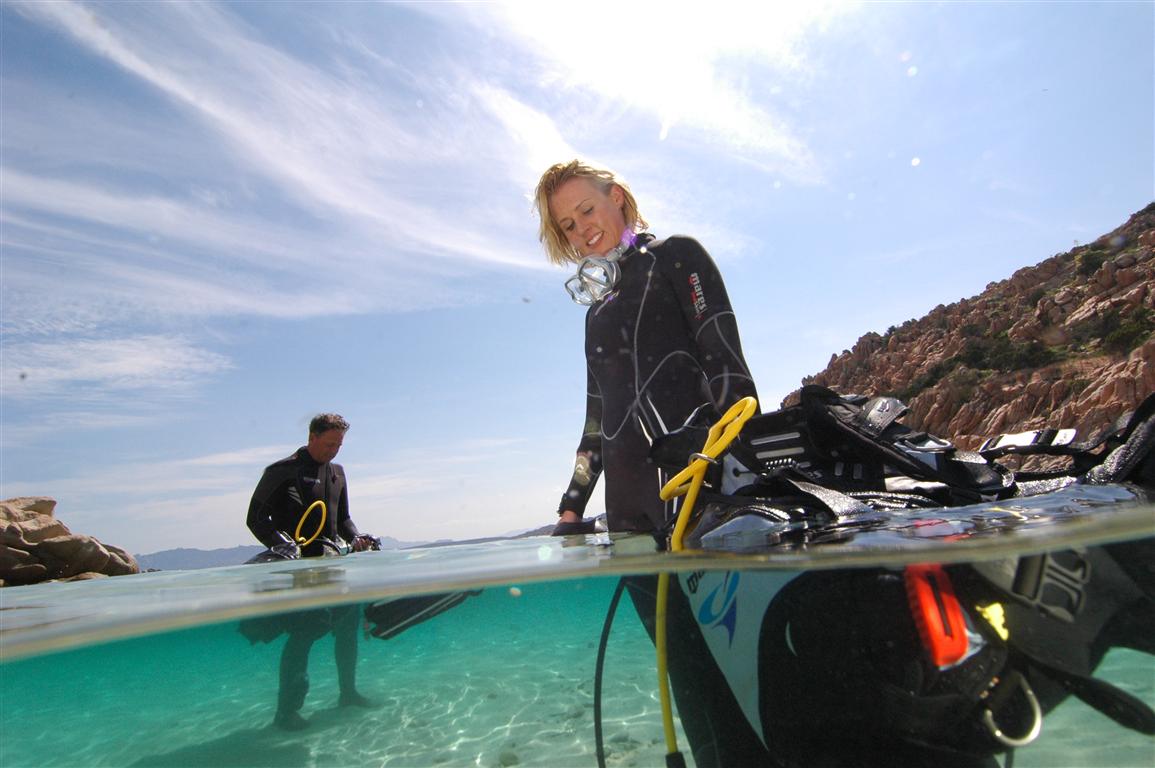 Deux plongeurs se préparant à une plongée sous-marine dans les eaux cristallines de Hyères, avec équipement de plongée et paysage côtier en arrière-plan.